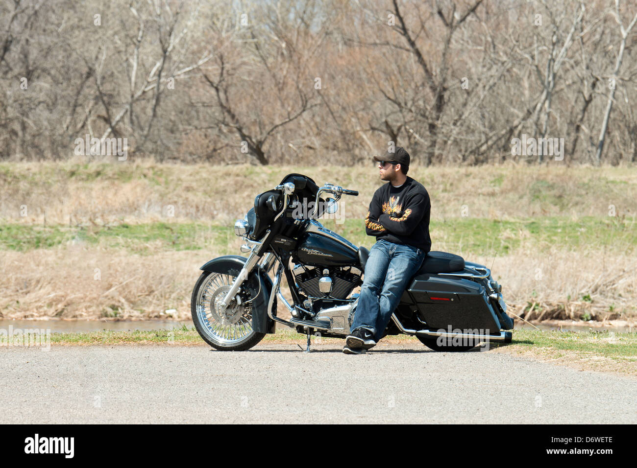 A man in his late 20s or early 30s, sits sideways on his Harley Davidson motorcycle and enjoys the early spring day. Oklahoma, USA. Stock Photo