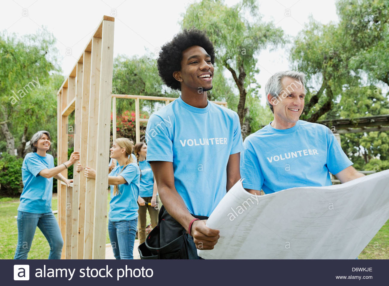 Men holding blueprint with volunteers constructing wooden frame in background Stock Photo