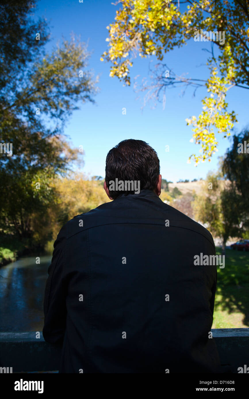 Back view of a man, deep in thought, leaning on a bridge in a rural setting. Stock Photo