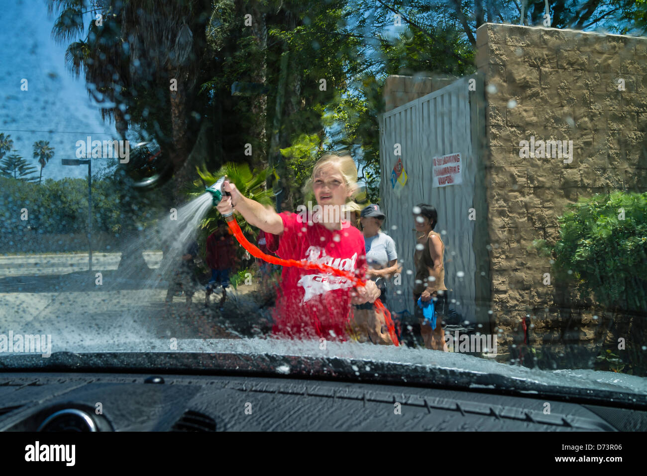 A female teenage student uses a hoses to rinse off a car at a high school fund-raising car wash as viewed from inside the car. Stock Photo