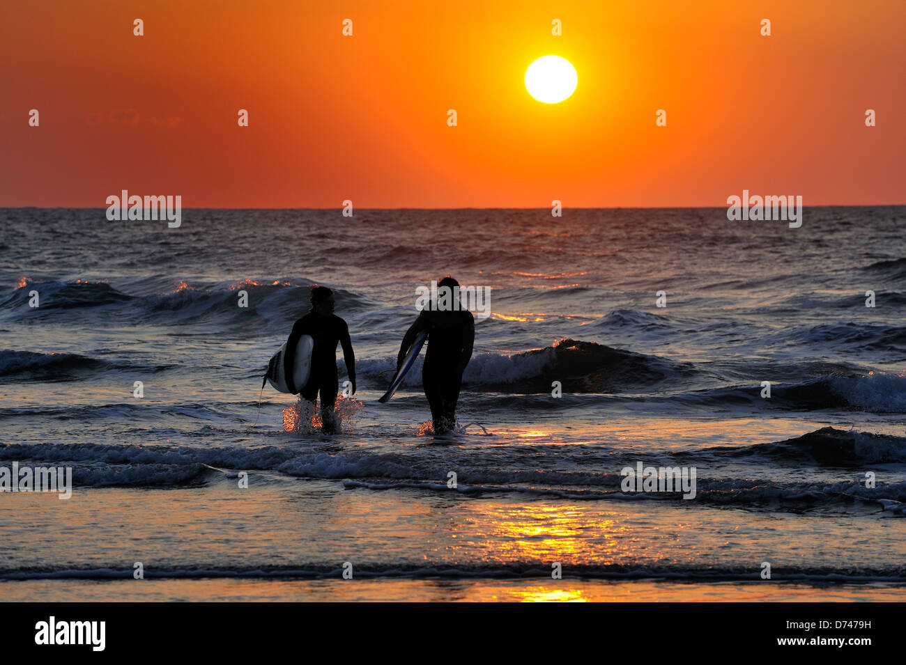 The sun sets behind two surfers leaving the sea after a day surfing Stock Photo