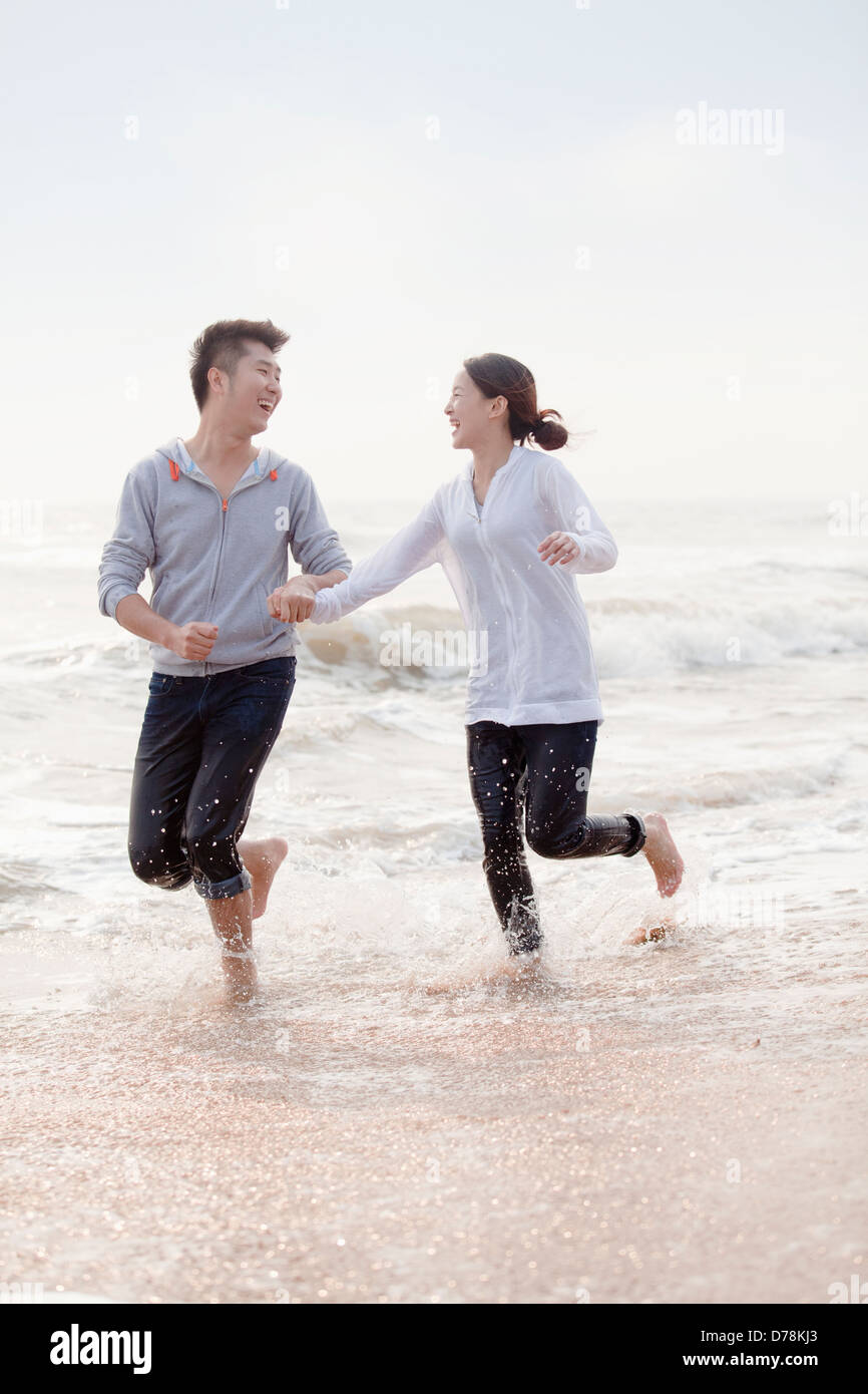 Young couple holding hands and running by the waters edge on the beach ...