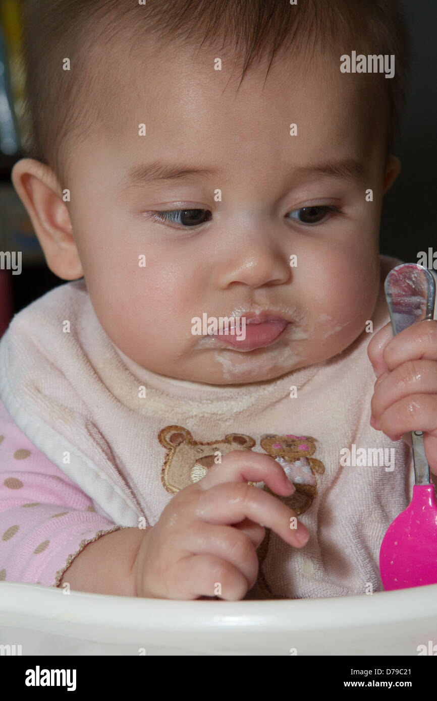 Unimpressed five-month-old baby reacts to first solid food as she holds her cereal spoon. Stock Photo