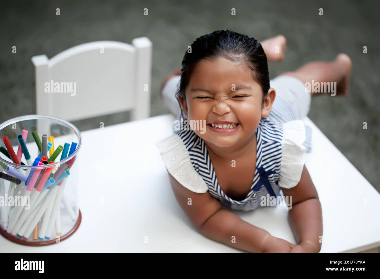 Little girl with cute smile laying on top of a white kiddie table with colorful markers Stock Photo