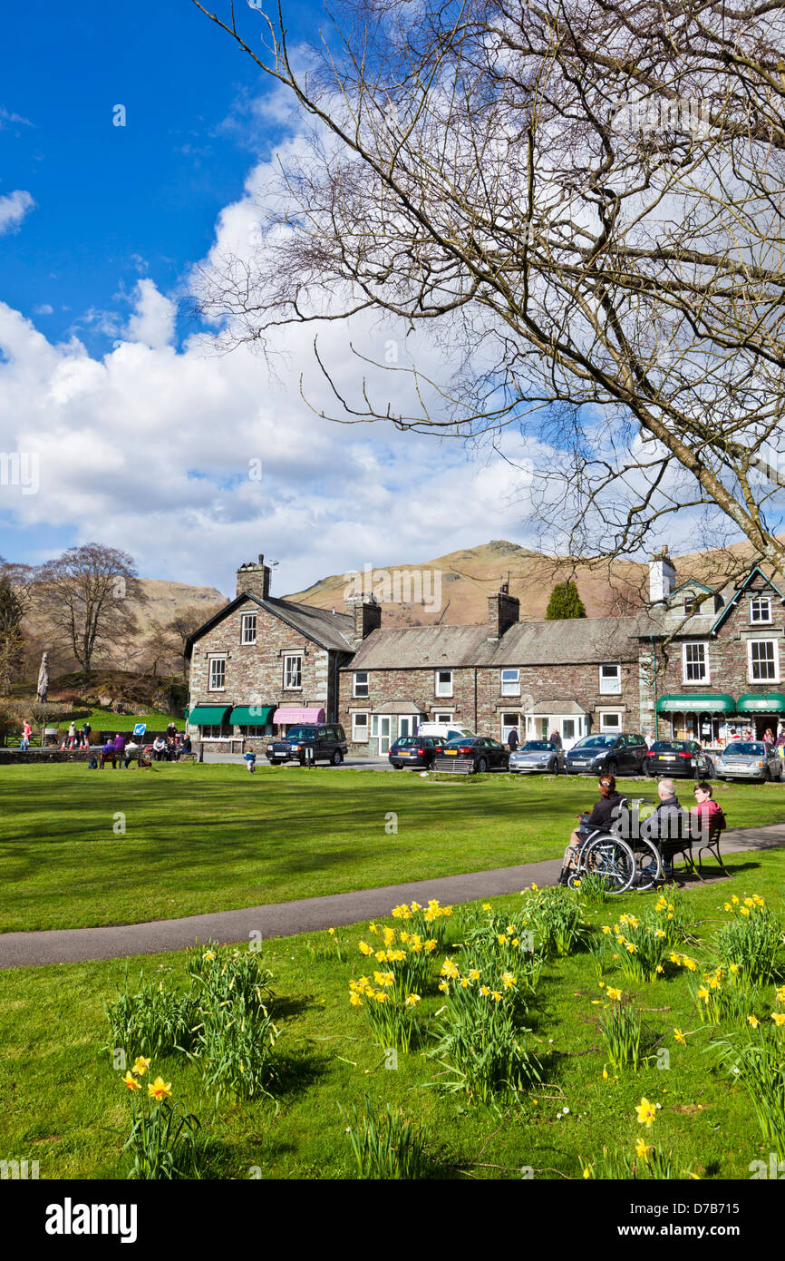 People sat on benches around the village green enjoying the spring sunshine  in Grasmere Cumbria England UK GB EU Europe Stock Photo