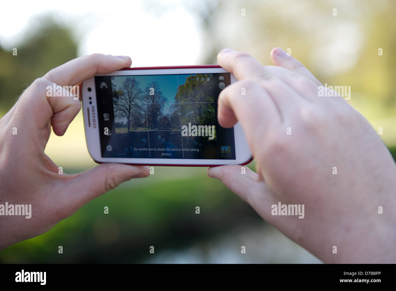 Hands are seen holding a Samsung Galaxy S3 phone, taking a photo of a park scene on the camera app of a river in a park. Stock Photo