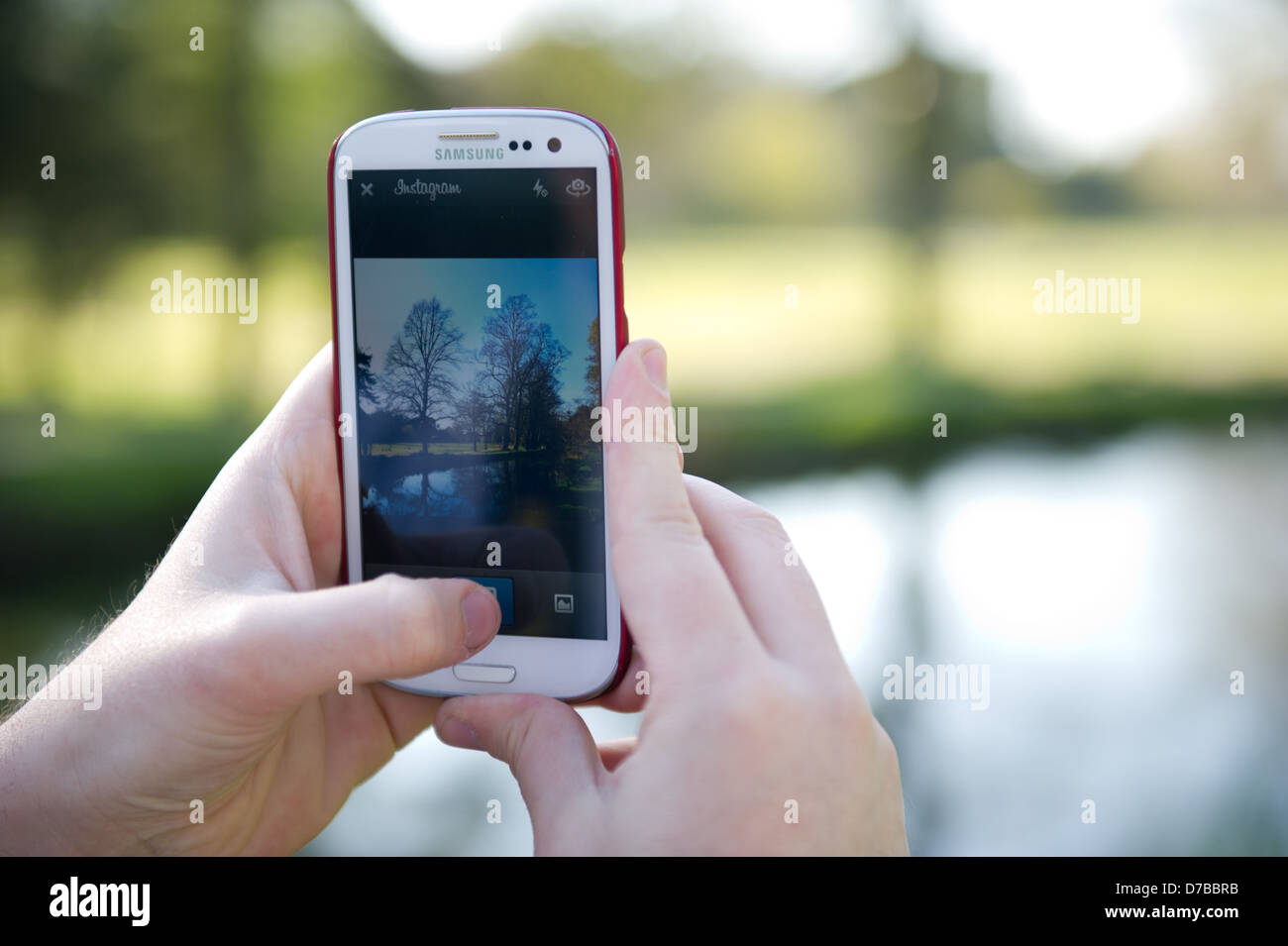 Hands are seen holding a Samsung Galaxy S3 phone, taking a photo of a park scene on the Instagram app of a river in a park. Stock Photo