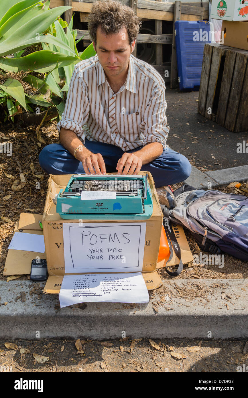A 25-30 year old male sits on the ground with his Smith-Corona typewriter on a cardboard box selling original poems. Stock Photo