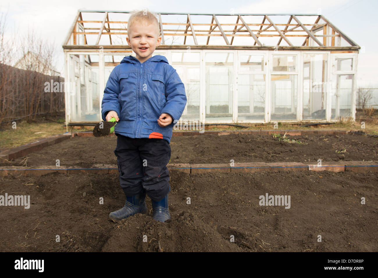 Happy smiling joyful child (boy) digging soil, working in spring garden in village. Little farmer. Stock Photo