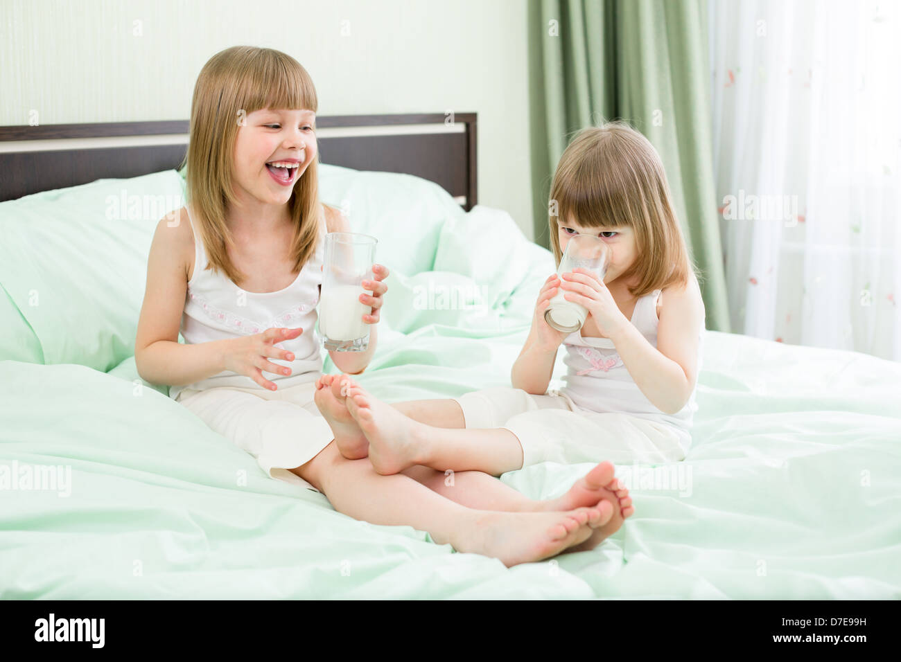 Two little girls drinking milk from glasses on bed Stock Photo