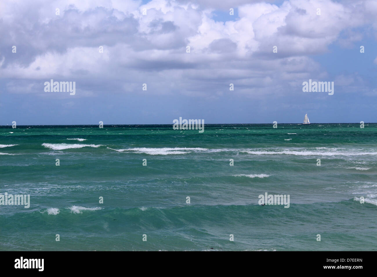 Lone sailboat with full sails on the horizon of the ocean with choppy waves making their way to the shore and expressive skies. Stock Photo
