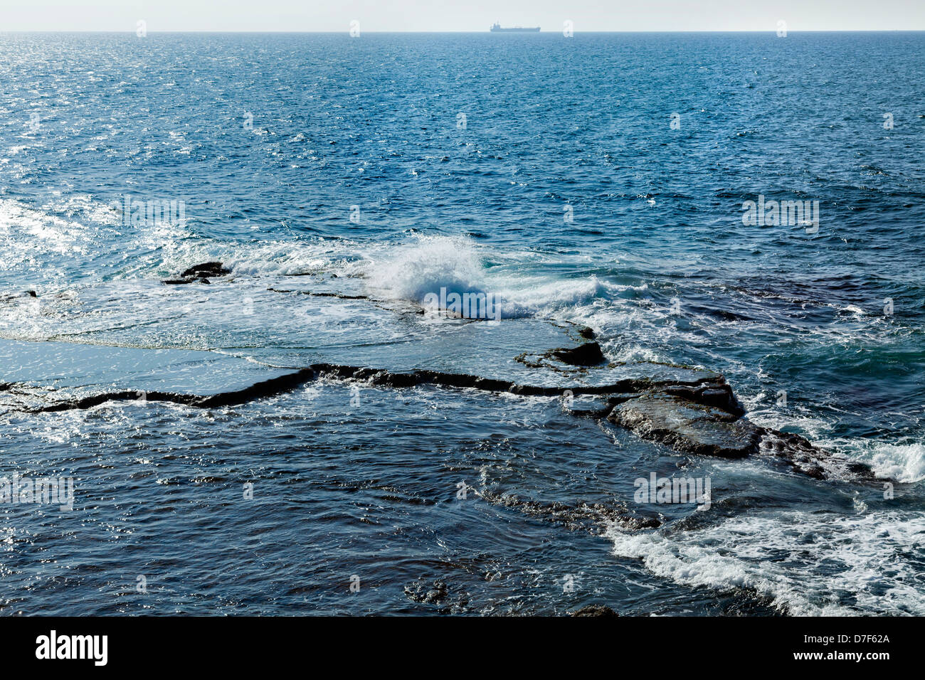 Waves crashing against a large flat rock in the Mediterranean sea. A large freighter breaking the horizon. Stock Photo