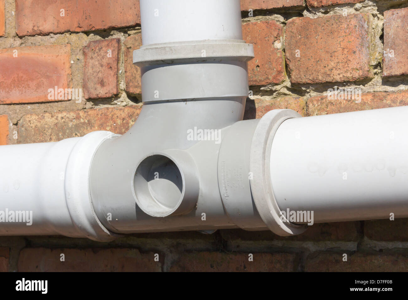 Grey plastic (Polyvinyl Chloride, un-plasticised - PVC-U) drainpipe T piece section, mounted horizontally on a brick wall. Stock Photo