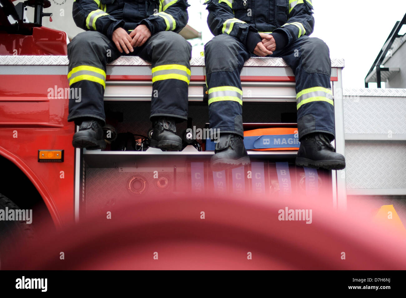 Two firemen sitting on a fire truck. In the foreground is a hose. Photo