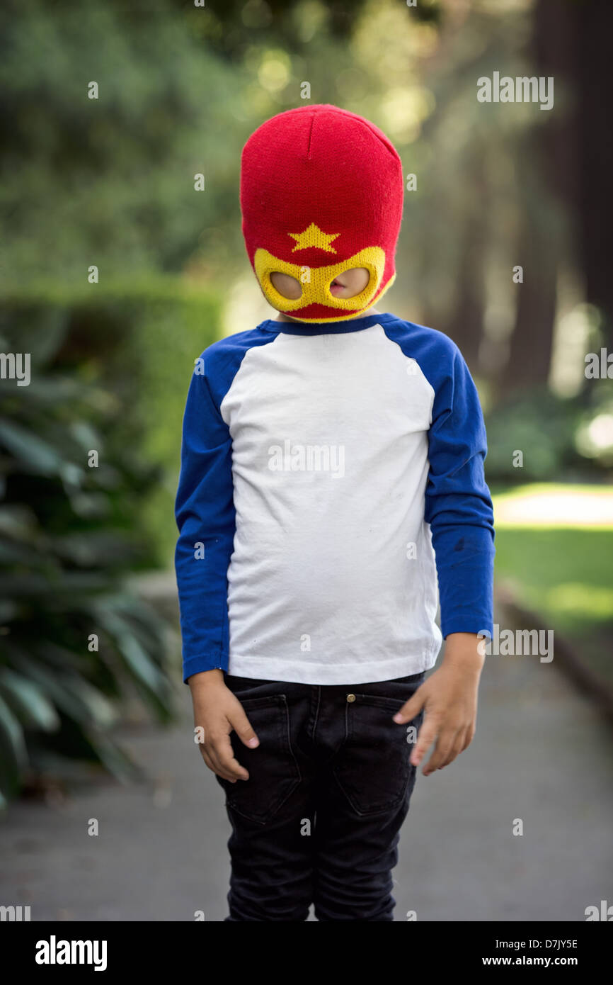 little boy wearing red and yellow superhero mask posing outside on the sidewalk, obscuring his eyes Stock Photo