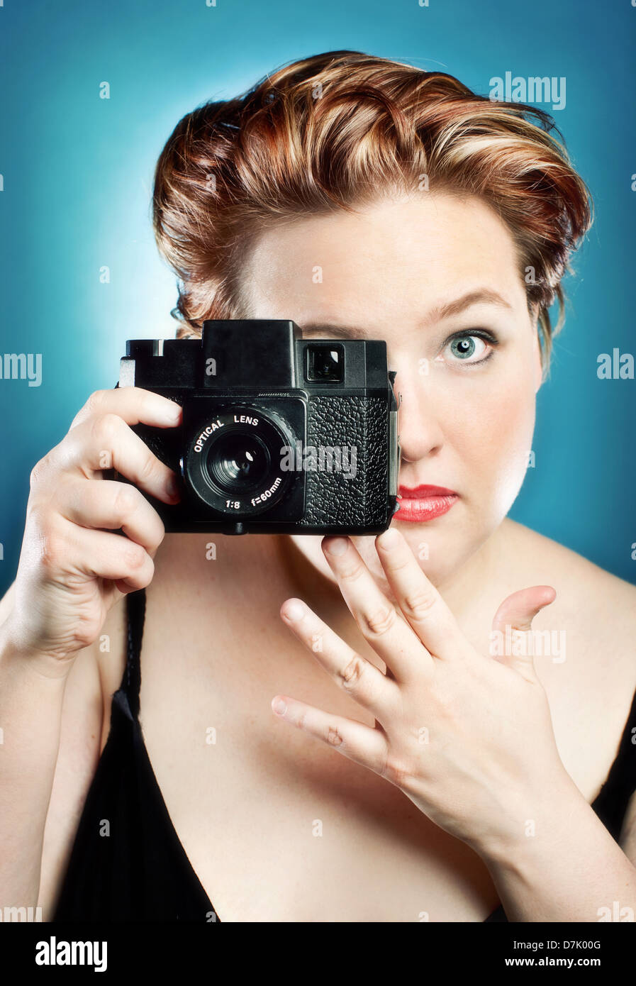 Portrait of young woman taking a picture with a toy camera in studio against blue backdrop Stock Photo