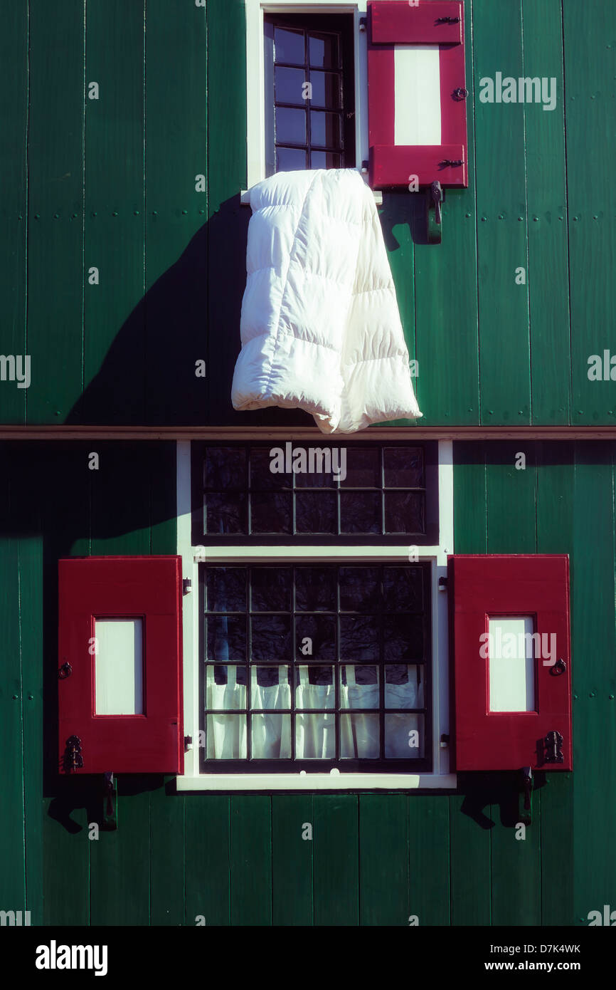 a white duvet hanging out of a window of a green wooden house with red window shutters Stock Photo