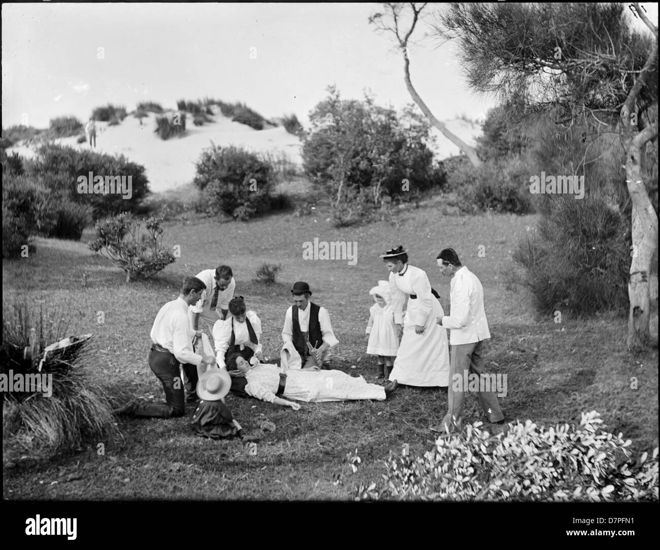 The image shows a woman unconscious on the ground, captured in a black-and-white photograph. The scene is staged in an outdoor setting, possibly near a picnic, with children and family members nearby. The setting includes natural elements like sand dunes, trees, and shrubs, suggesting a summer environment. The image evokes themes of concern and emergency, with fainted subjects and possibly staged moments from a fight or accident. Stock Photo