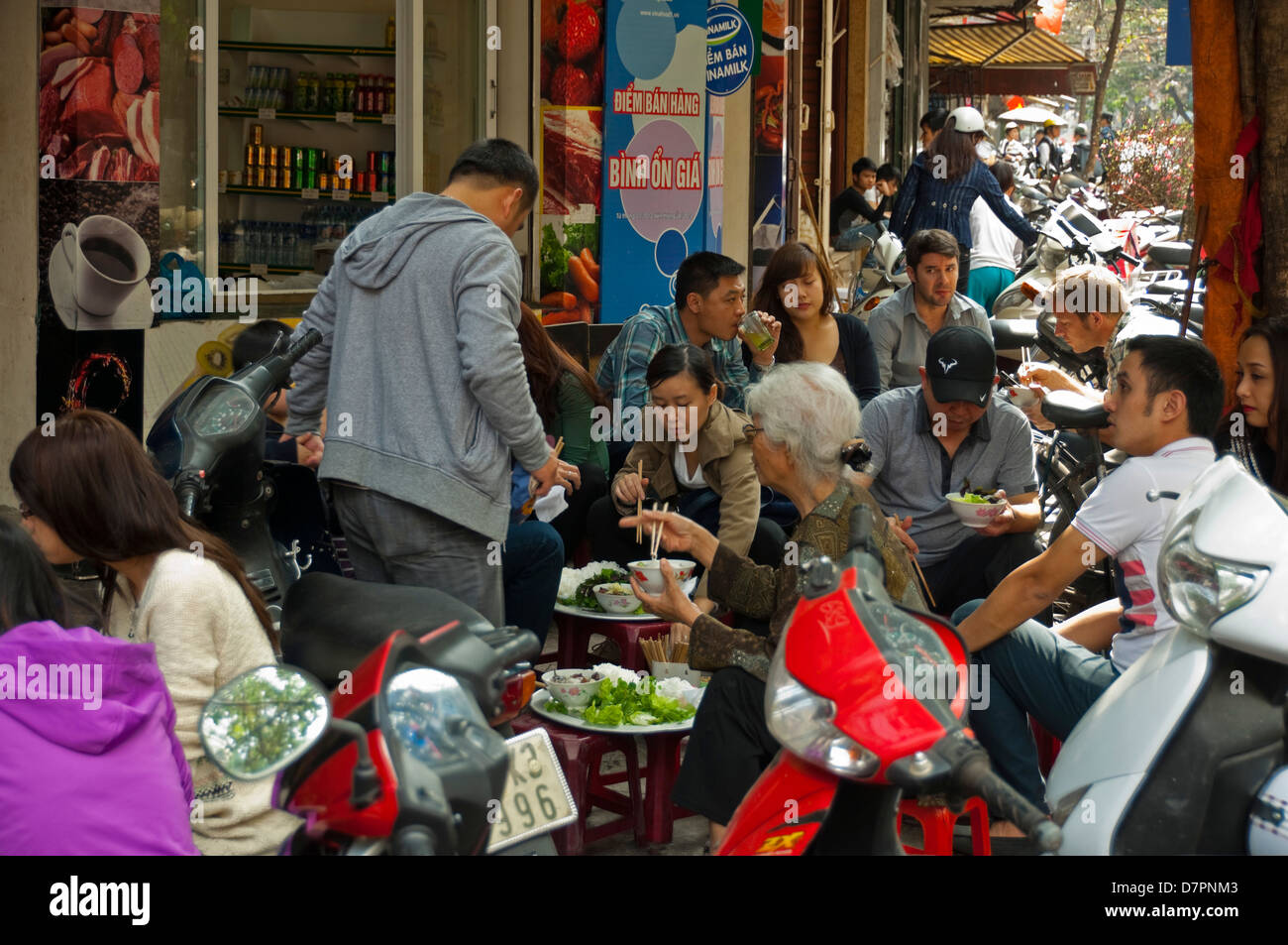 Horizontal view of people sitting in the street eating traditional Pho, noodles, on the pavement in Hanoi. Stock Photo