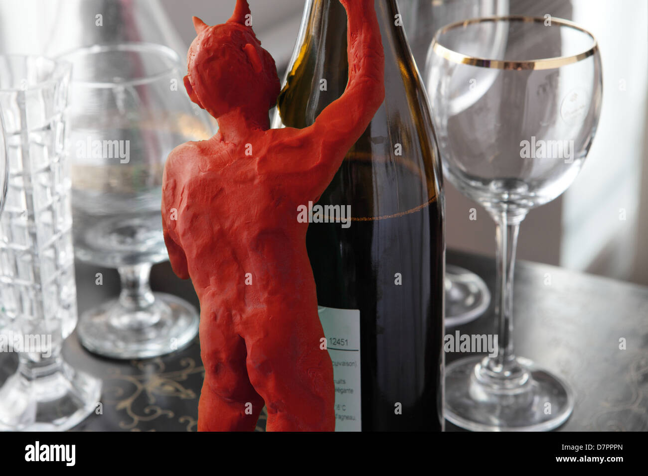 temptatia little red devil, (modelling clay) seen from the back, hides himself behind a bottle of wine. Some glasses are spread out on an inlaid table Stock Photo