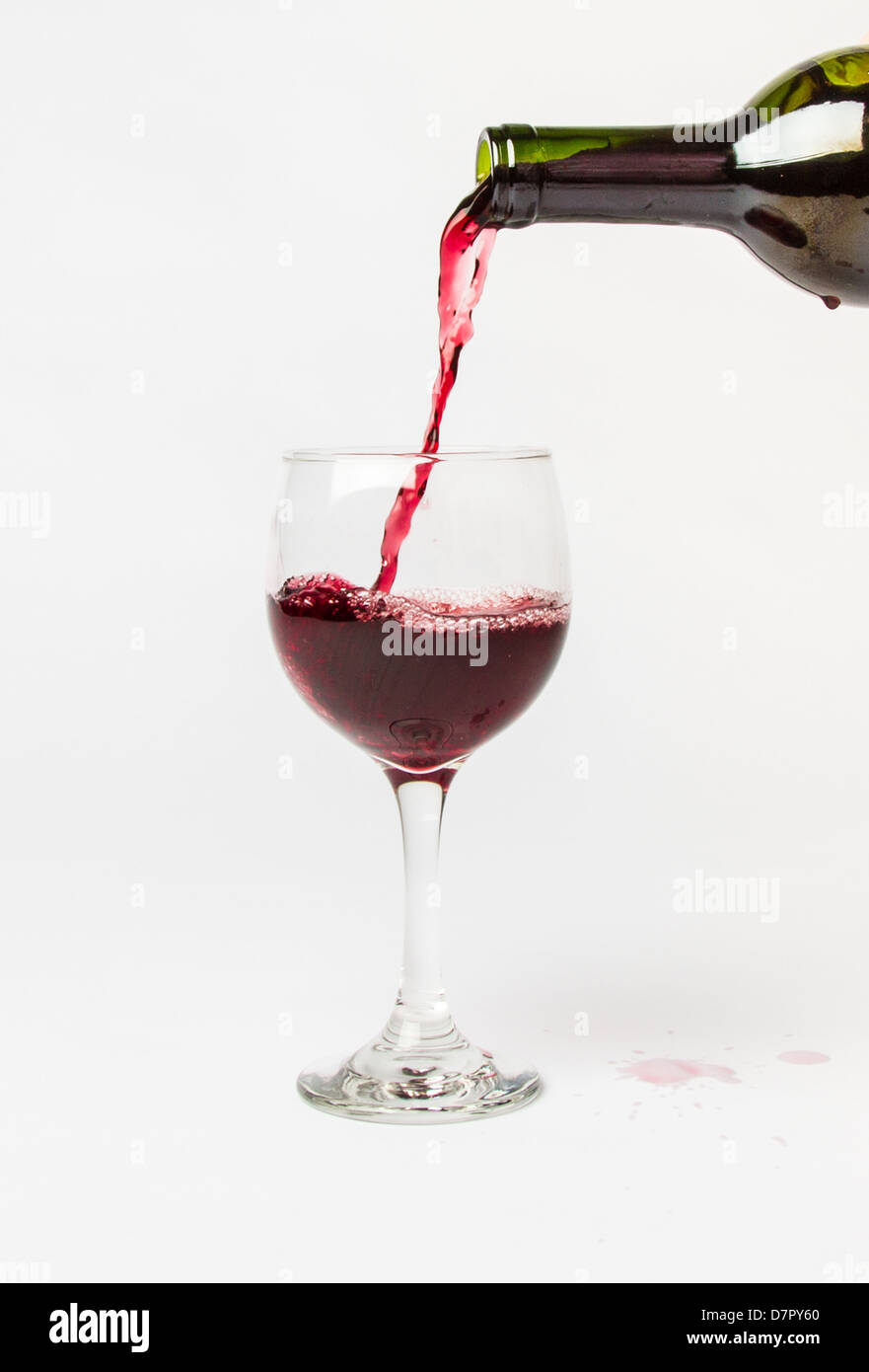 Red Wine pouring out of a bottle into a glass and splattering the white background. Stock Photo