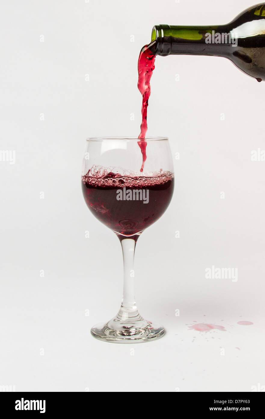 Red Wine pouring out of a bottle into a glass and splattering the white background. Stock Photo