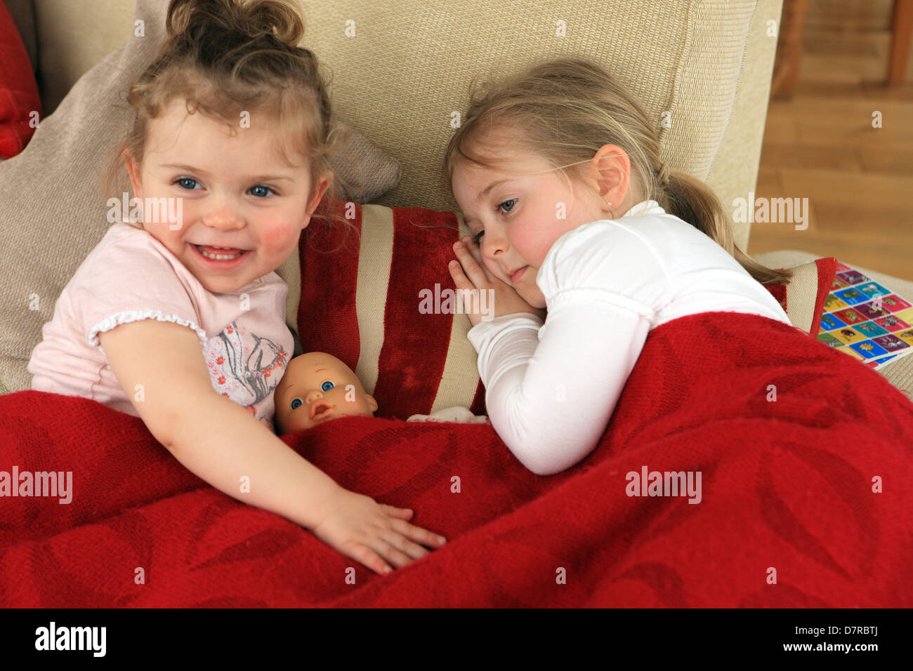 Sisters lying in a makeshift bed on the couch along with their doll. One sister is not feeling well. Stock Photo