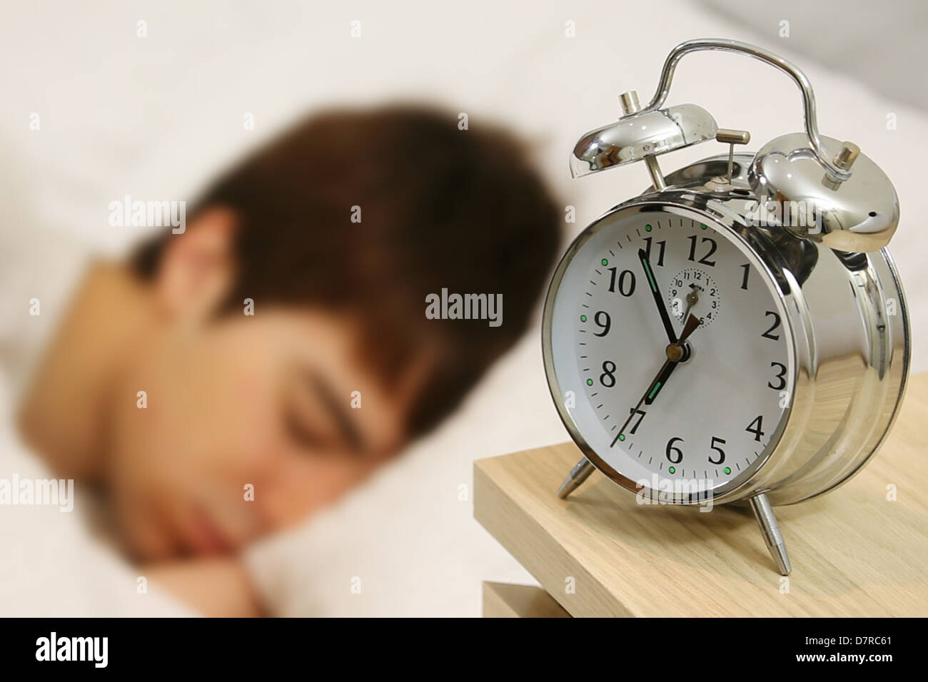 Young male adult asleep in bed, an alarm clock by his bed just before 7am. The alarm is just about to ring. Stock Photo