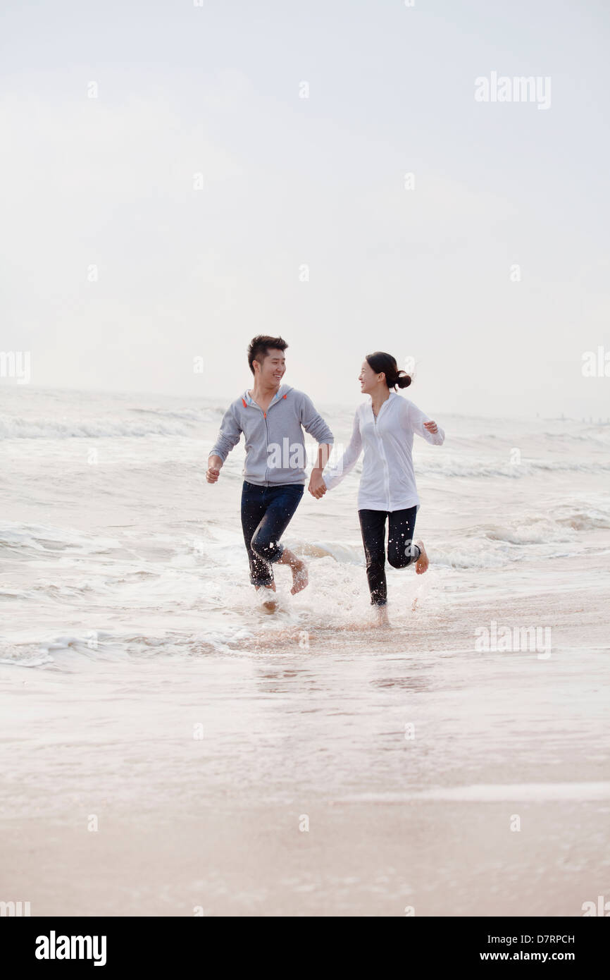 Young couple holding hands running by the waters edge on the beach ...