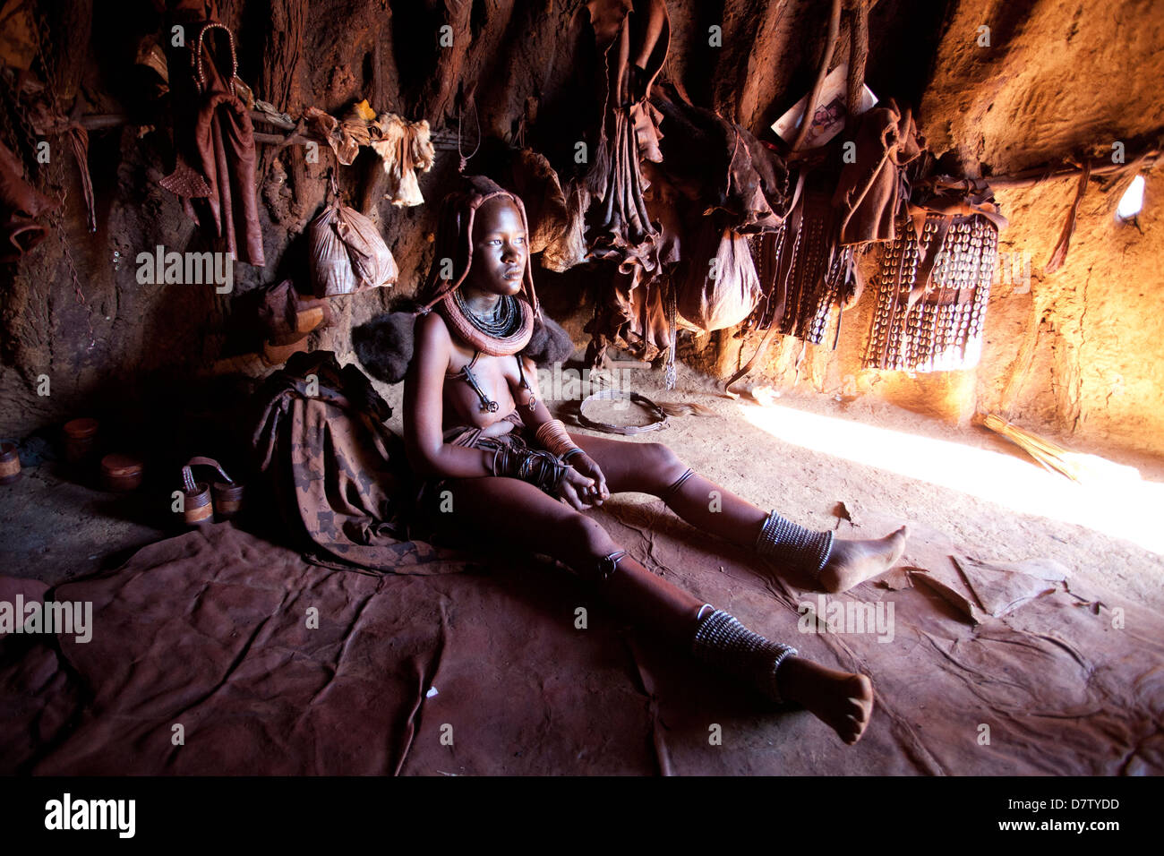 Himba woman inside a mud dwelling hut wearing dress and jewellery and covered in Otjize, Kunene Region, Kaokoland, Namibia Stock Photo