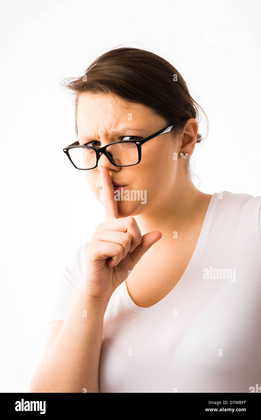 A strict young brunette teenage Caucasian girl wearing glasses with her finger to her lips, calling for silence Stock Photo