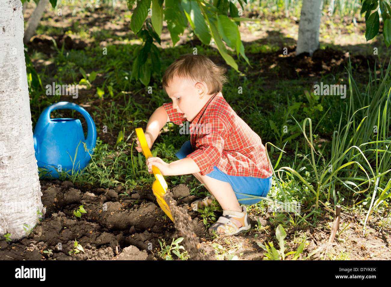 Little boy digging in the garden at the foot of a tree with his colourful yellow toy spade Stock Photo