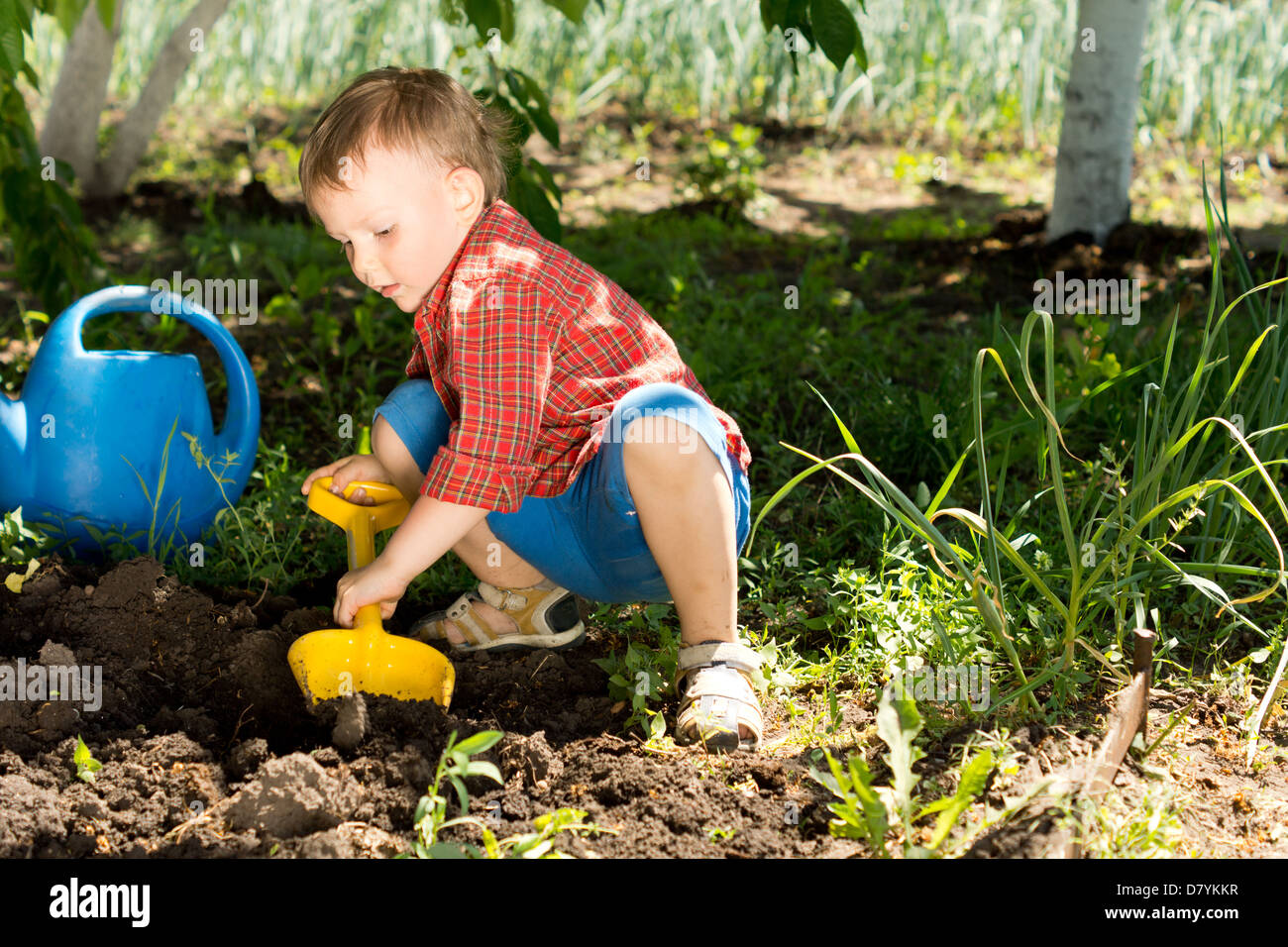 Little boy digging with a colourful yellow toy spade in fresh earth under the shade of a tree in the vegetable garden Stock Photo