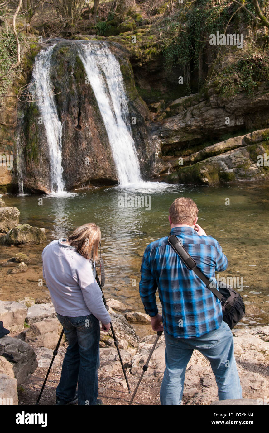 Rear view of 2 photographers using tripods (man & woman) taking photos of scenic waterfall - Janet's Foss, Malham, Yorkshire Dales, England, UK. Stock Photo