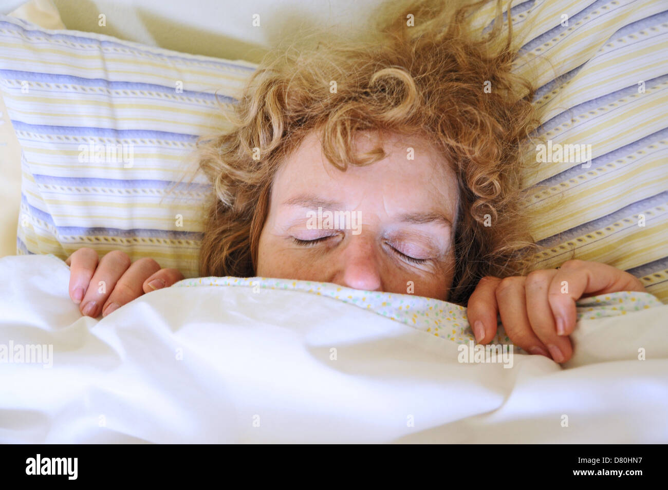 woman 50s lying asleep under the bed covers with top of head and eyes poking out Stock Photo