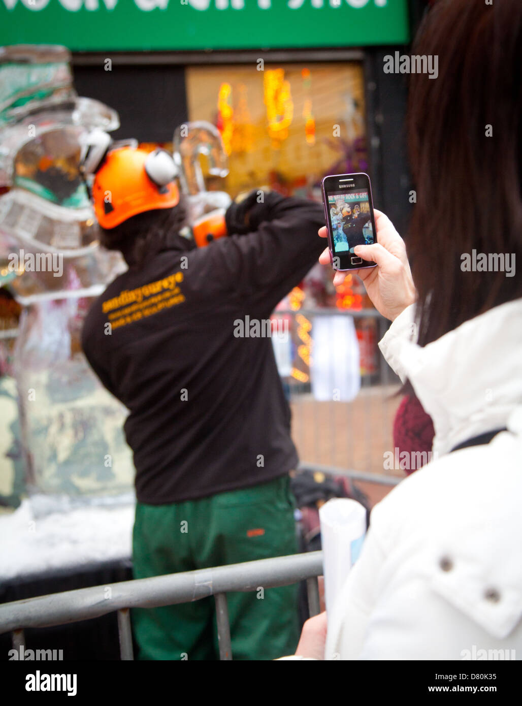Woman taking iphone photos of two men carving a block of ice into a Snowman Sculpture on Rhyl town centre high street Stock Photo