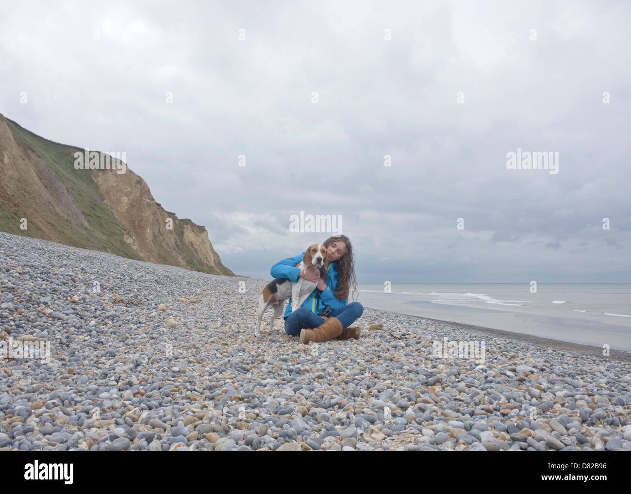 Woman sitting cross-legged on pebble beach fussing dog. Cliffs and sea in background. Stock Photo