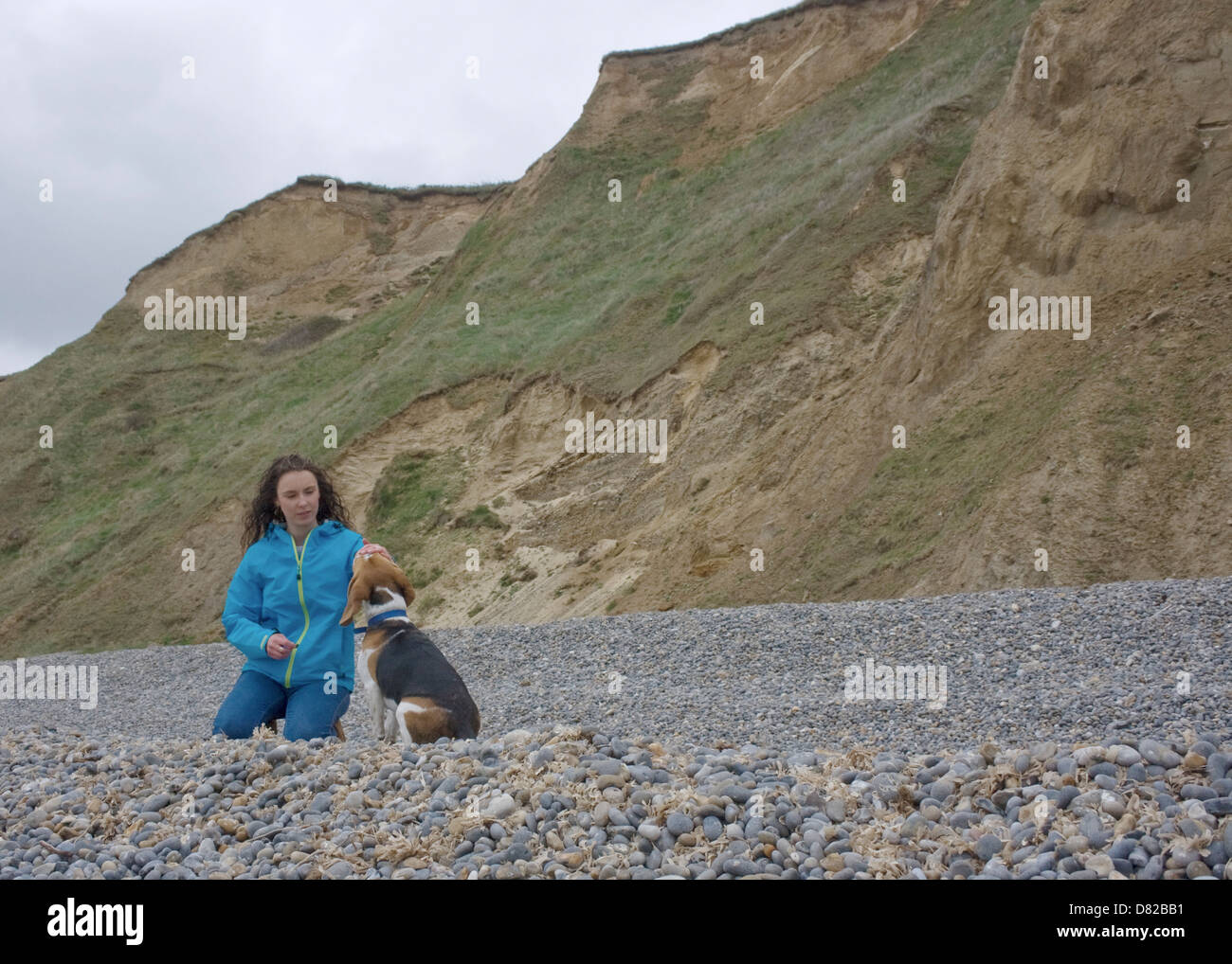 Woman kneeling on beach with cliffs in background fussing her dog. Stock Photo
