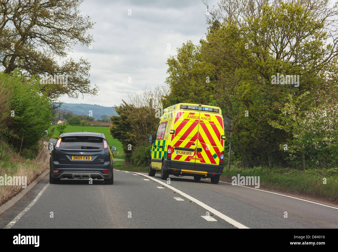 View through a windscreen of an ambulance with blue lights flashing rushing to an emergency overtaking a car along A5 road. UK Stock Photo