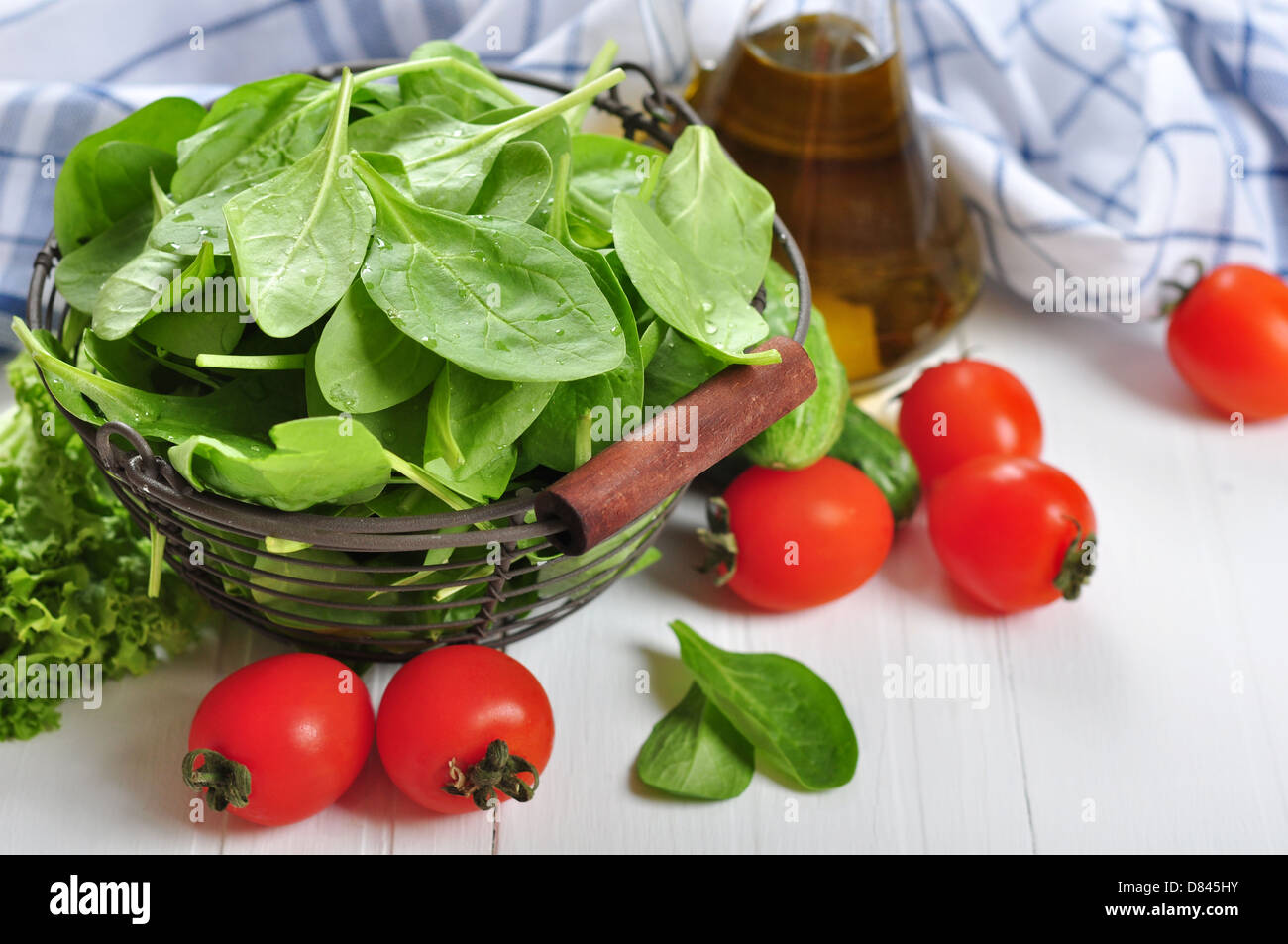 Fresh spinach leaves in basket with tomatoes, cucumber and olive oil on a white wooden background Stock Photo