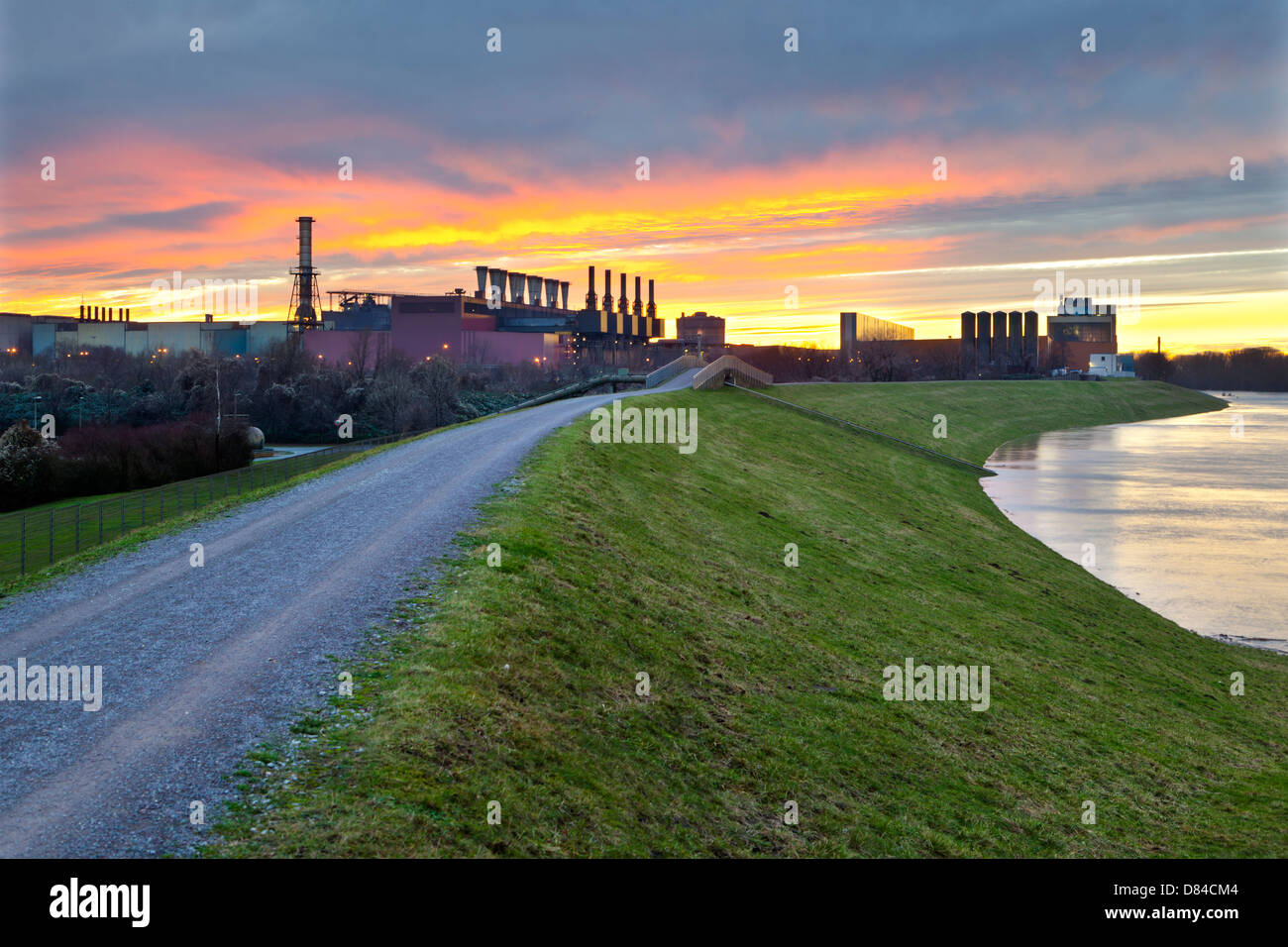 Steel plant next to river with a fiery evening sky, a dyke in the foreground. Stock Photo