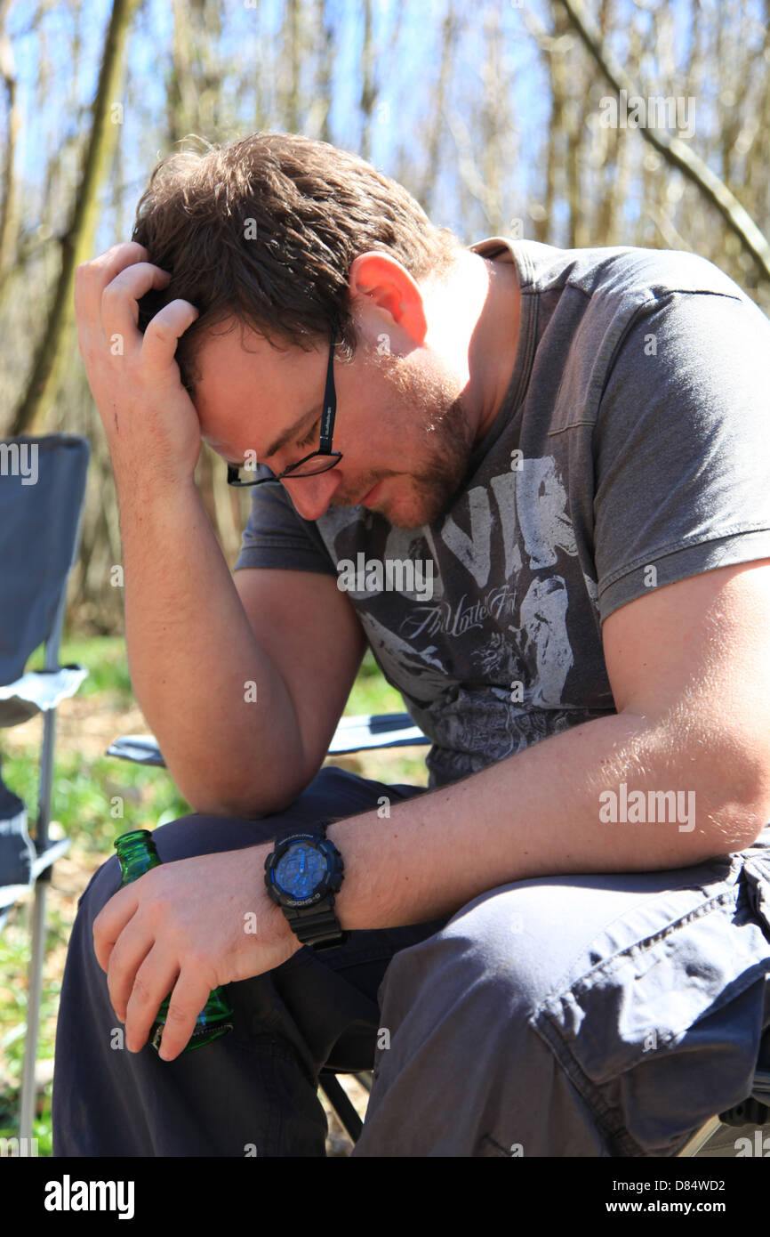 Man in his early 30's drinking beer and looking fed up while camping in the woods. Stock Photo