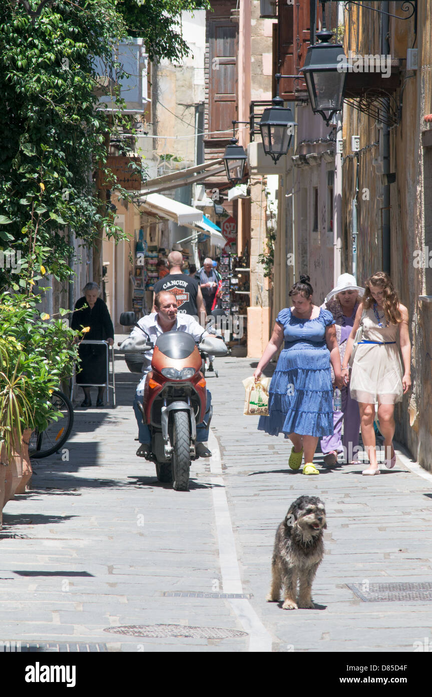 A man rides a motorbike past a group of women and a dog within a narrow street  within the old town of Rethymno, Crete Stock Photo