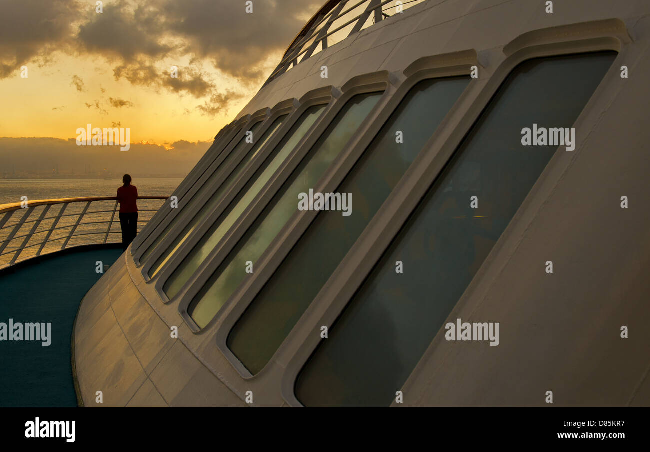 A woman standing on the deck of a cruise ship and watching a sunset ...