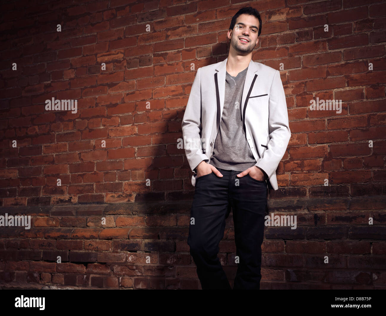 Portrait of a fashionable smiling young man wearing light gray blazer and black jeans standing at a brick wall Stock Photo