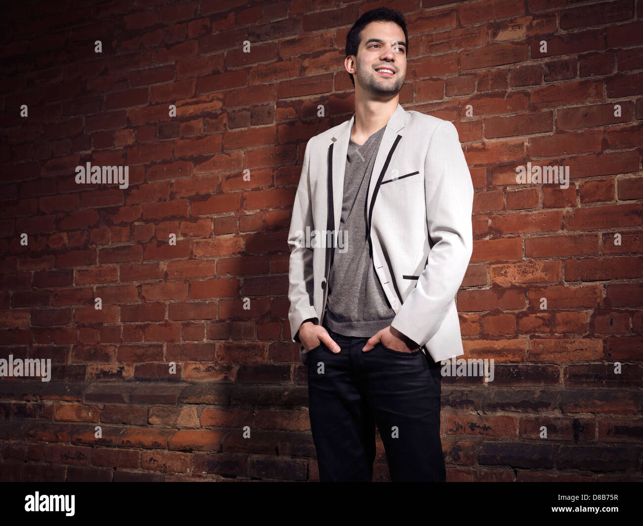 Artistic portrait of a smiling young man wearing light gray blazer and black jeans standing at a brick wall Stock Photo