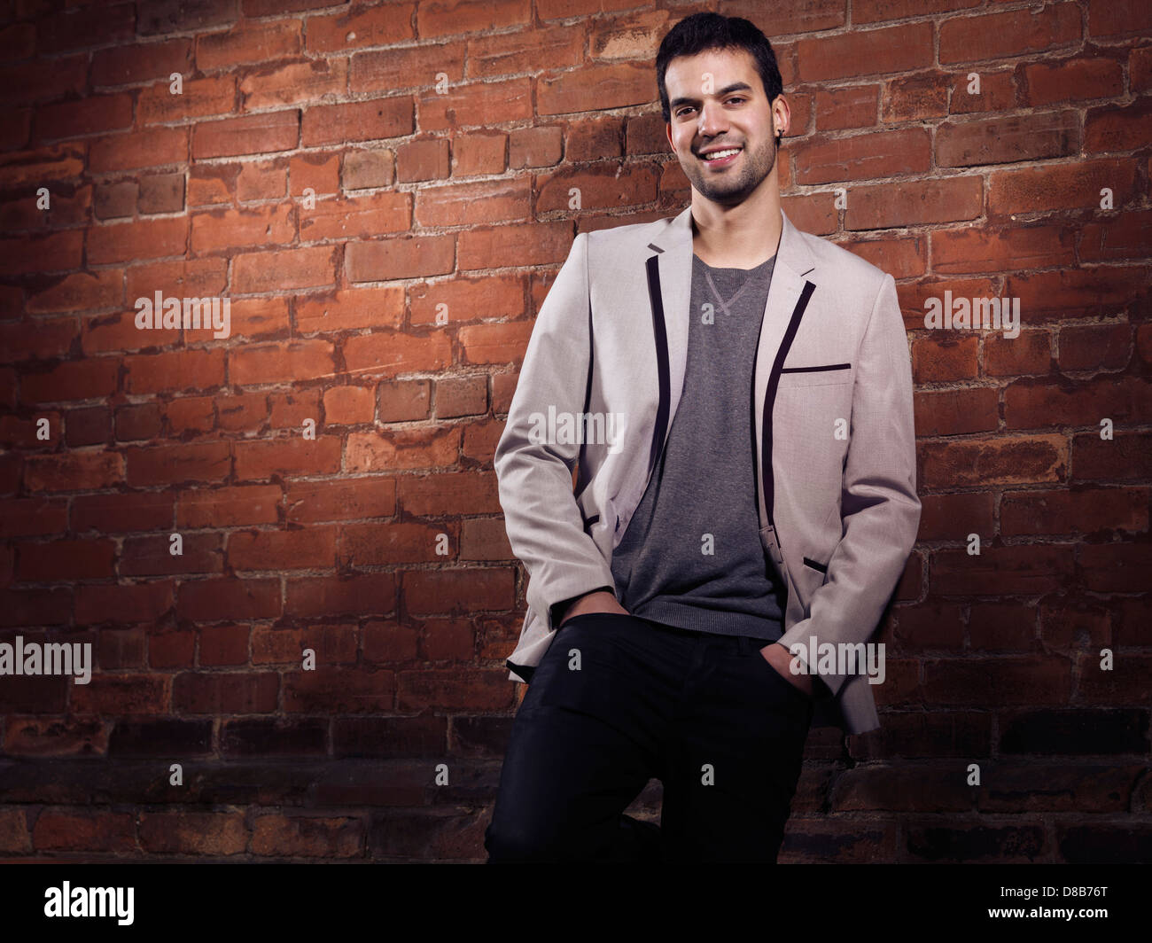 Artistic portrait of a smiling fashionably dressed young man wearing light gray blazer and black jeans standing at a brick wall Stock Photo