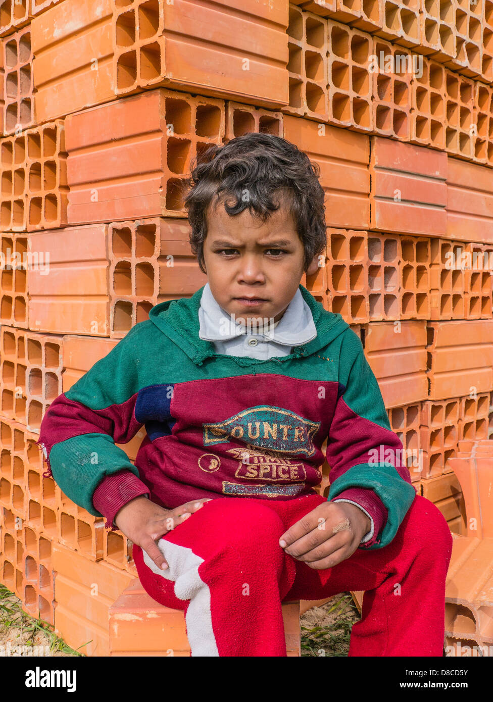 A young Hispanic boy sits in front of a pile of ceramic house building blocks at a Habitat for Humanity building site in Luque, Stock Photo