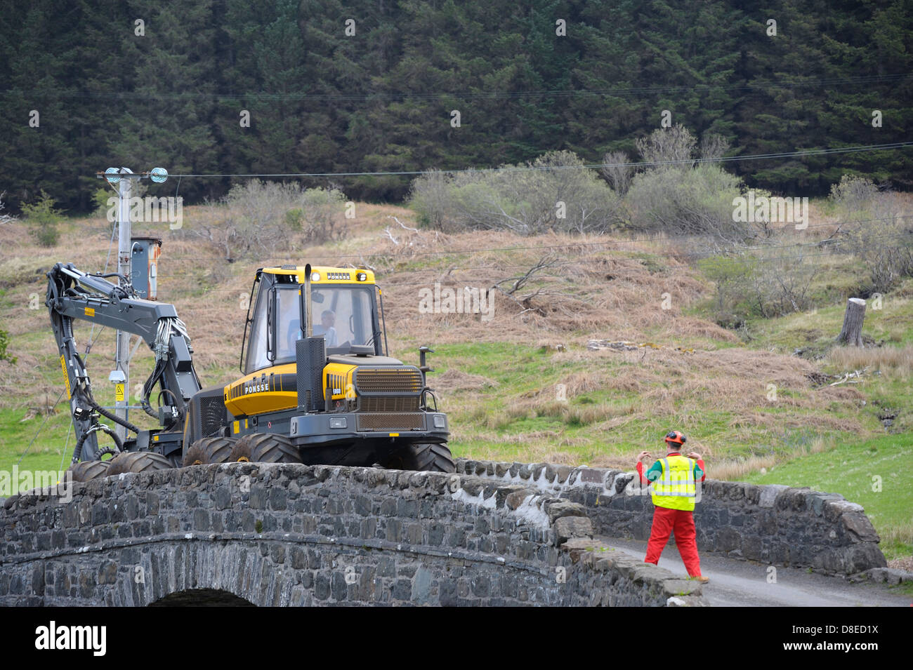 banksman directing large and heavy piece of timber harvesting machinery being moved through a small village on the isle of mull. Stock Photo