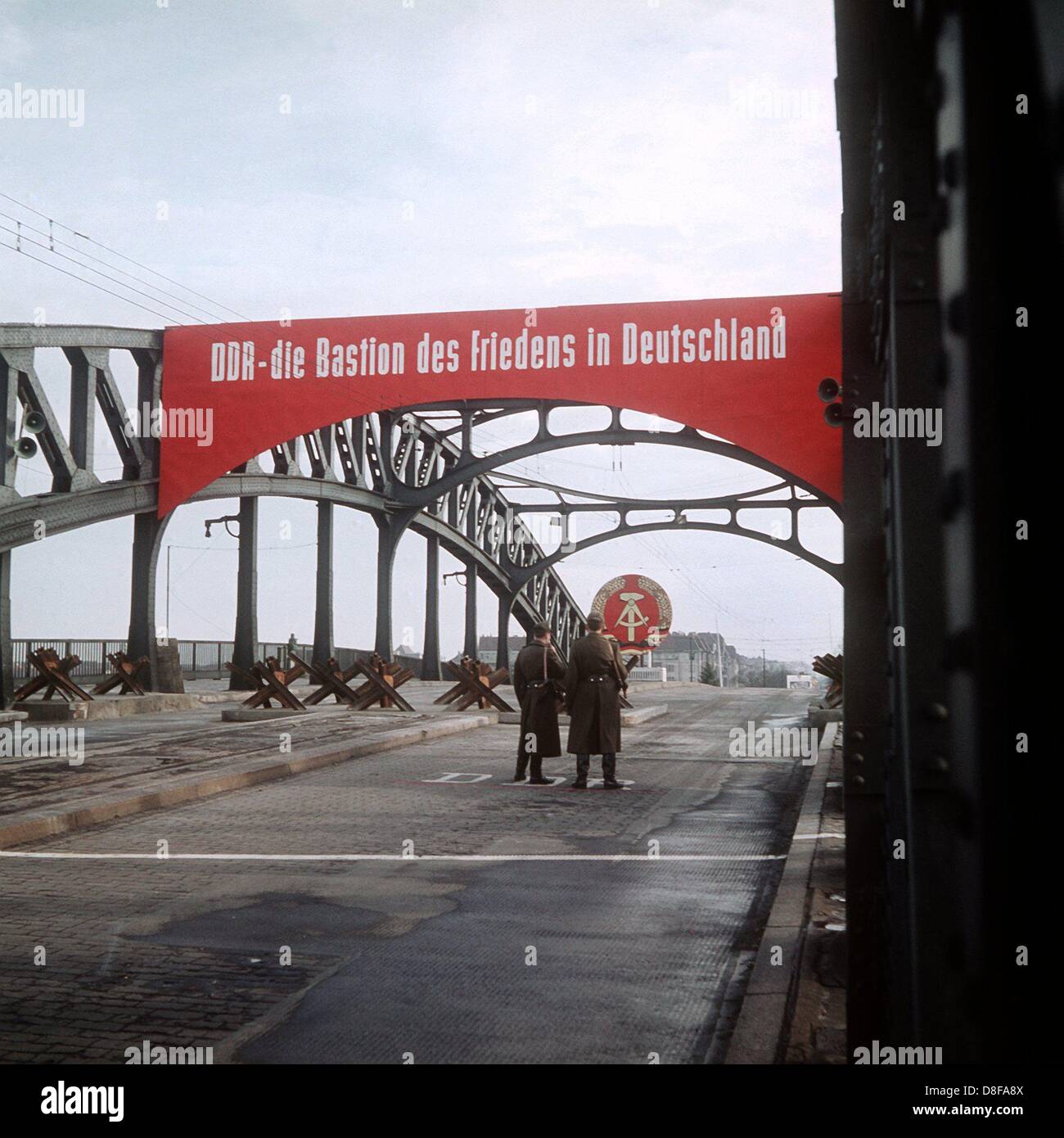 Unter einem Transparent mit der Losung:" DDR - die Bastion des Friedens in Deutschland" stehen zwei Volkspolizisten hinter Strassensperren aus zusammen geschweißten Straßenbahnschienen auf der Boesebrücke in der Bornholmer Straße im Norden Berlins. Der weiße Strich in der Mitte markiert die Grenze zwischen Ost- und Westberlin. Aufgenommen 1961. Stock Photo